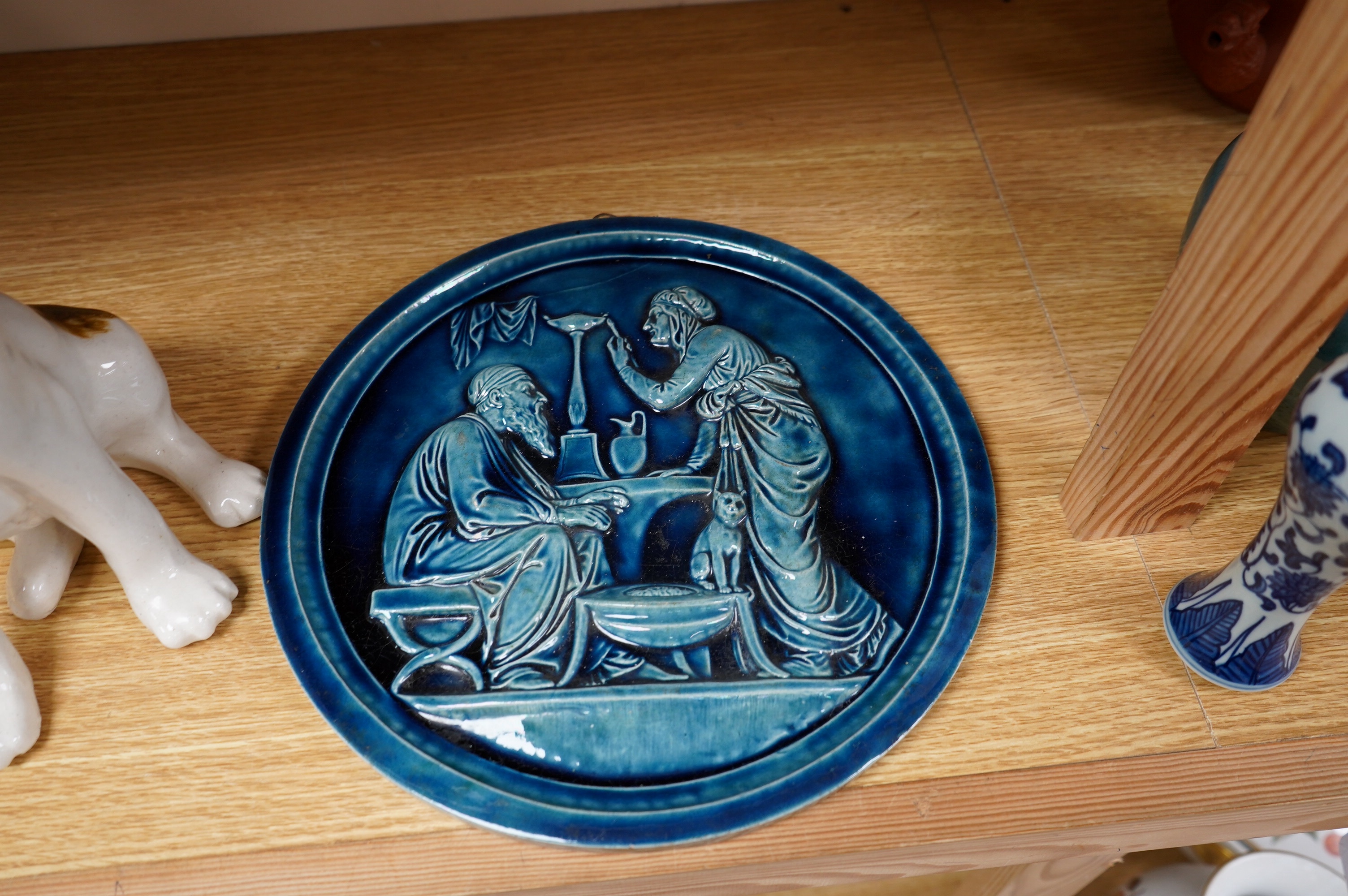 A Copenhagen P Ipson circular glazed plaque, representing Winter/Ages of Man, and a J. Winstanley pottery seated dog with glass eyes, largest 27cm in diameter. Condition - good, the right ear of the Winstanley dog has be